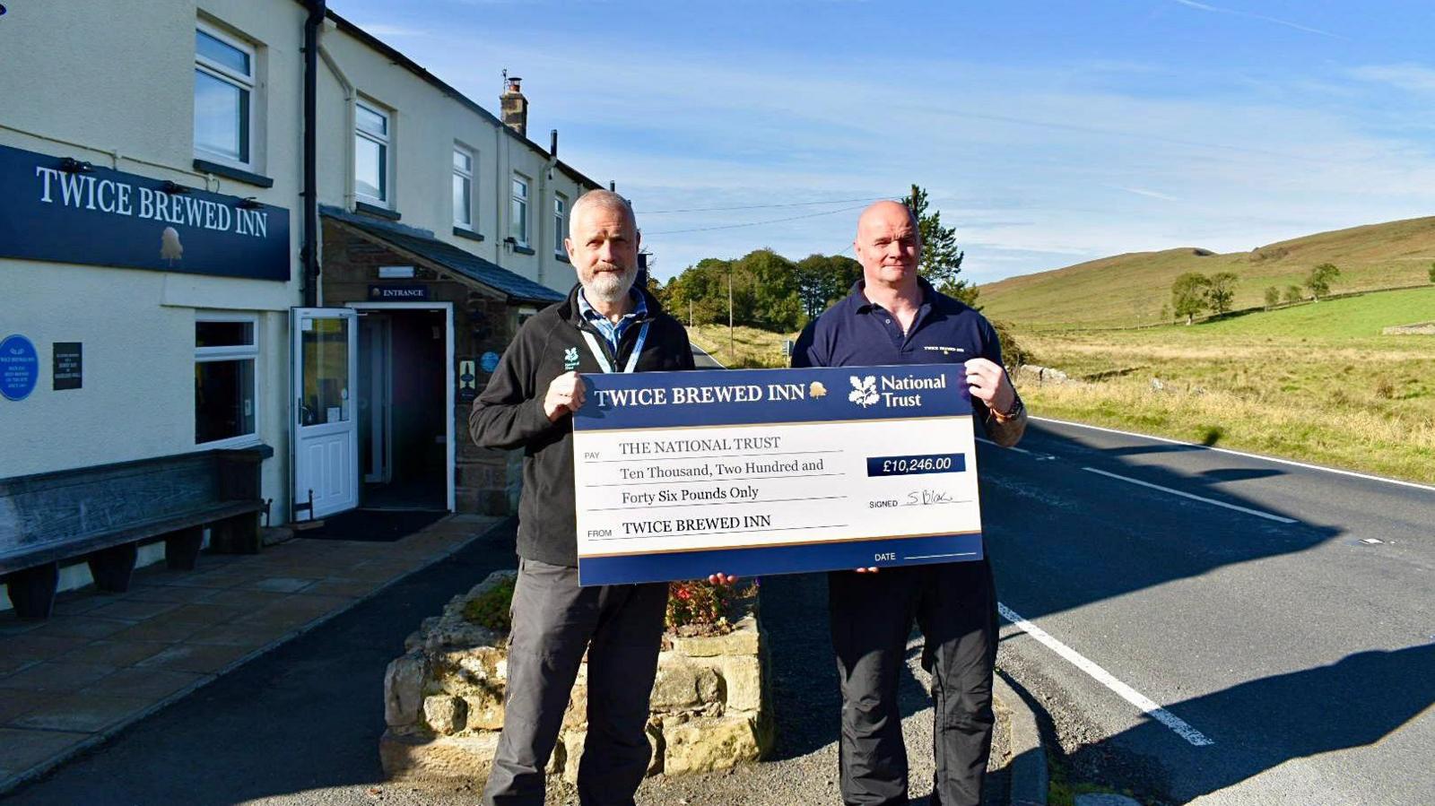Andrew Poad, from the National Trust, and pub manager Steven Blair. They are both wearing jeans and fleeces and holding up a charity cheque of £10,246 made out to the National Trust. They are standing outside the pub.