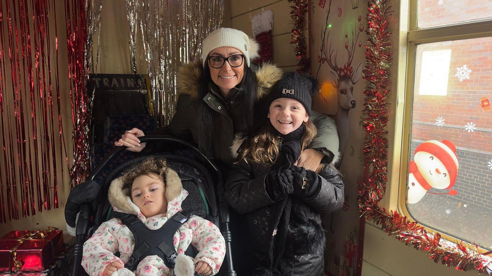 Tara and her daughters are in a carriage which has Christmas garlands and tinsel. There is a snowman decoration on the window and a picture of a reindeer. A Christmas present is wrapped up in shiny red paper.