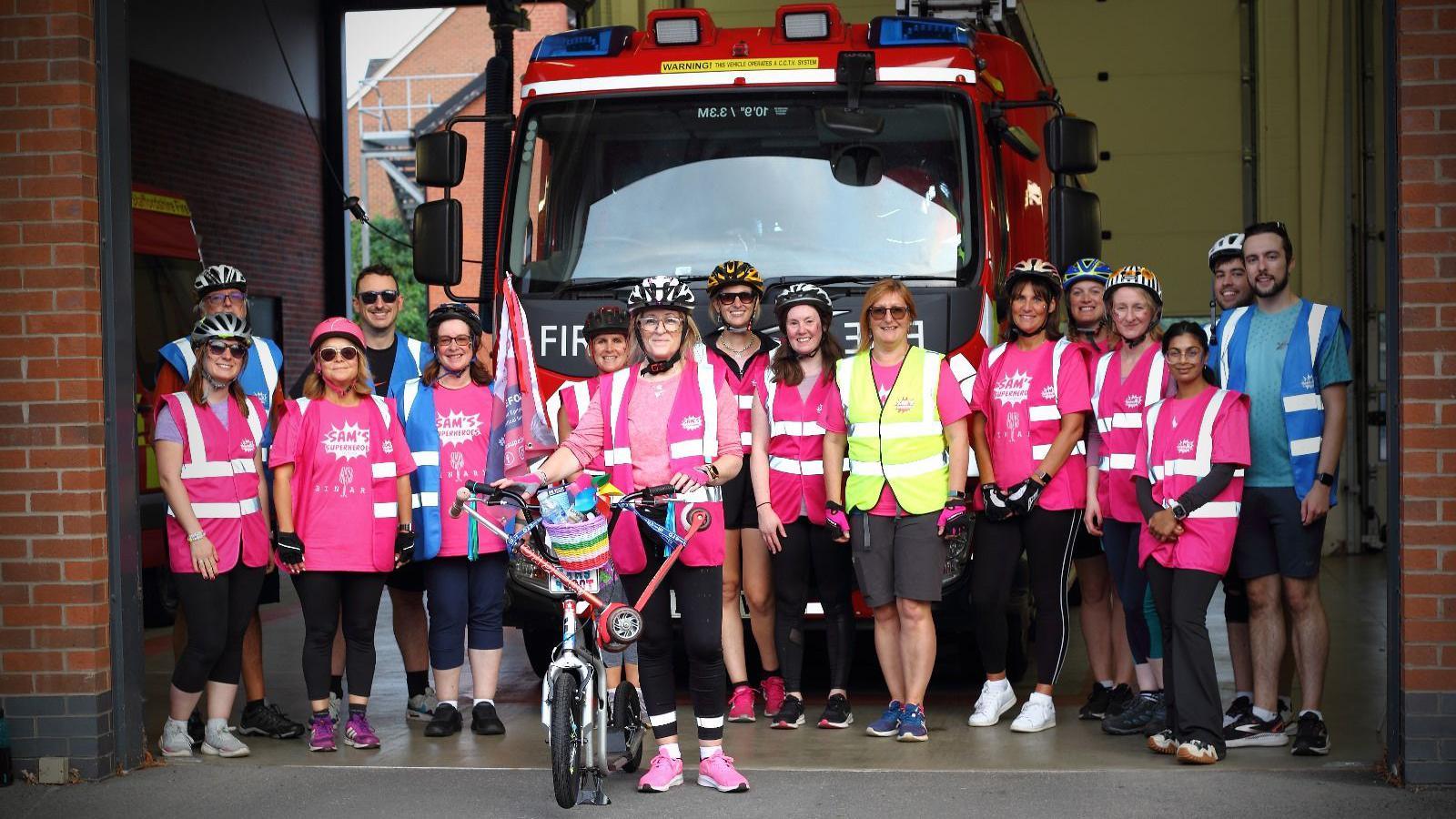 At the front of the photo is Rachel Liew on a scooter with a team of people stood behind her. They are at a fire station, and a fire engine is behind the group, who are all wearing either pink, blue or yellow hi vis jackets 