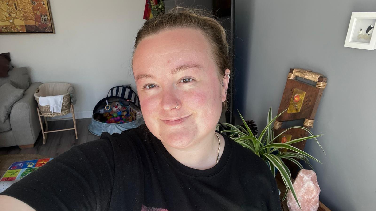 Anik has her dark brown hair tied back in a ponytail. She is wearing a black t-shirt and smiling directly at the camera from her living room.