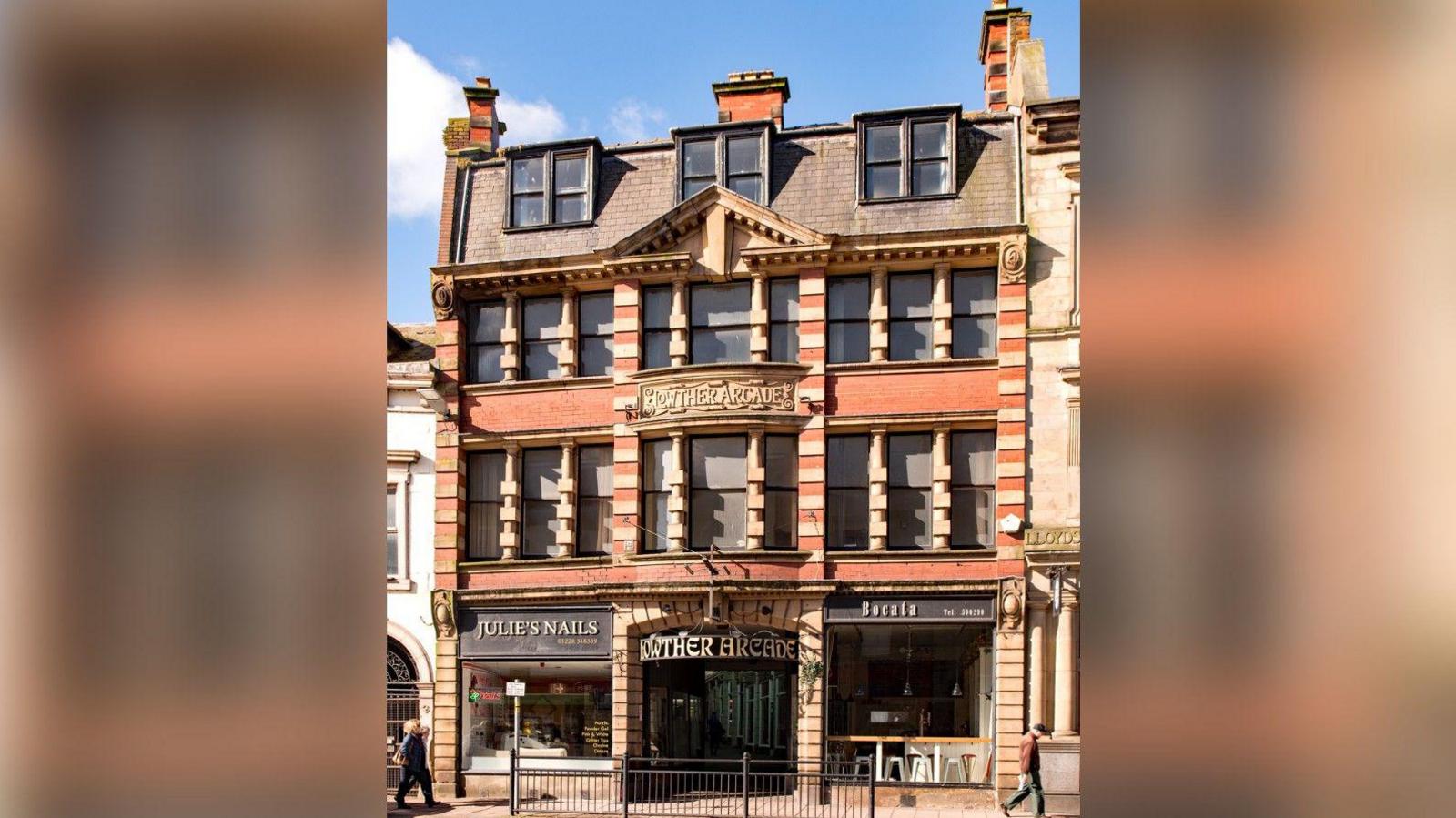 The entrance to the Lowther Arcade building in Carlisle, pictured in 2018. The four-storey building is in red brick with rusticated and alternate block quoins. There are two shops at the side of the entrance on the ground floor.