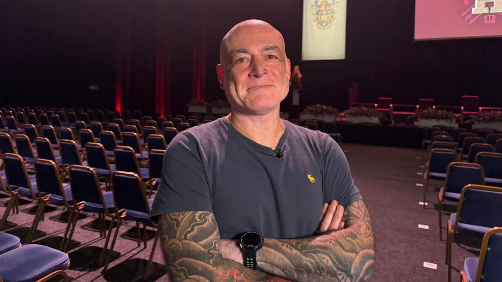 Dr Beardon has his arms crossed and smiles at the camera as he stands in a hall with several rows of chairs in front of a stage. He has tattoos on both his arms and is wearing a grey t-shirt and watch on his left wrist.