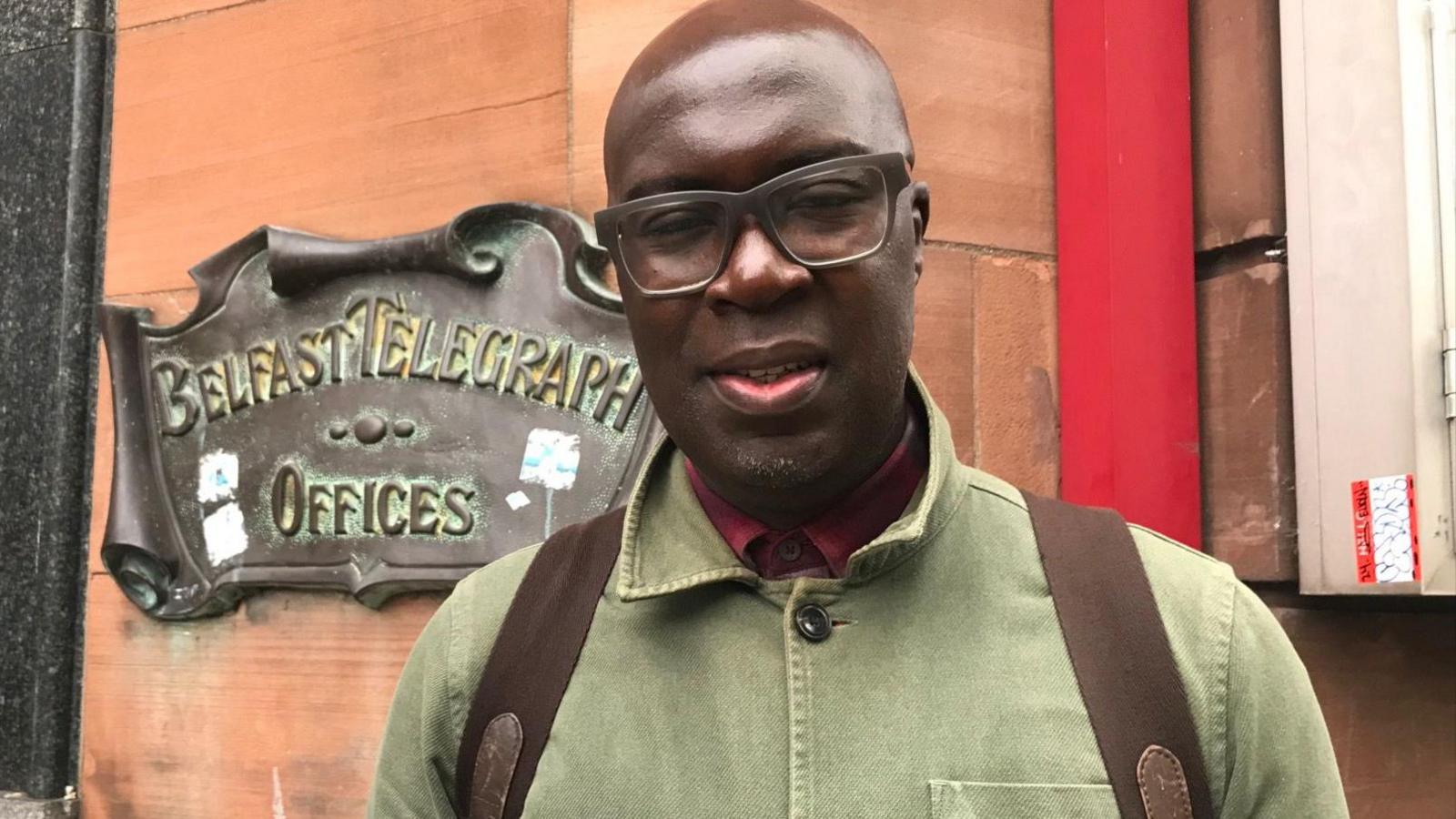 Kwame Daniels in front of the Telegraph building in Belfast.  He is wearing thick-rimmed glasses, a green jacket, a red shirt and a ruck sack.