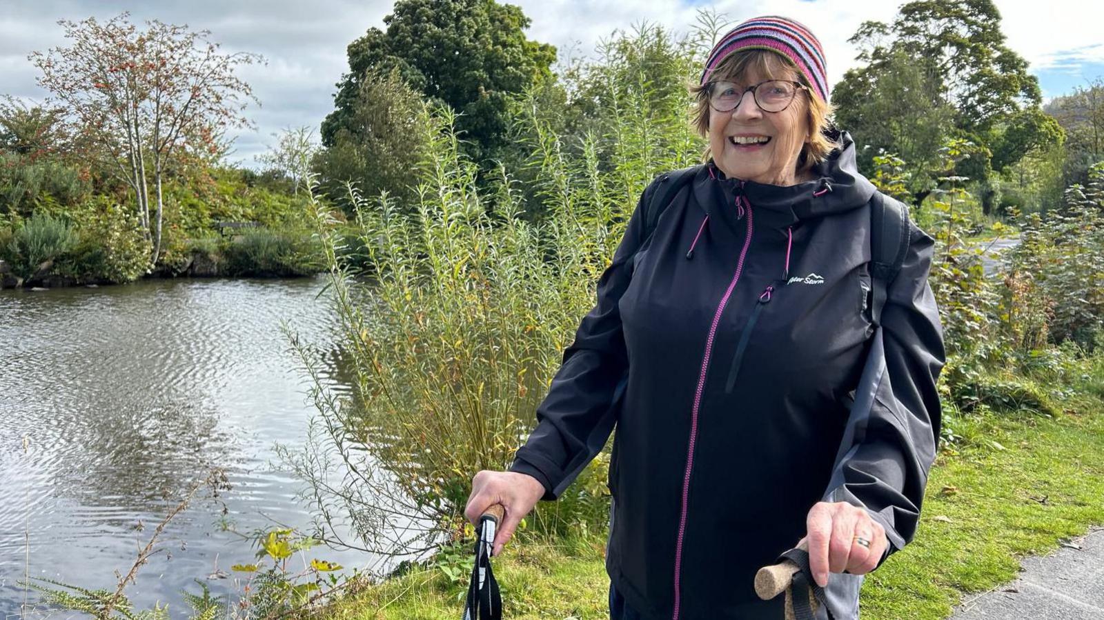 Sheila Mahon dressed in walking clothes with sticks, next to Ilkley Tarn.