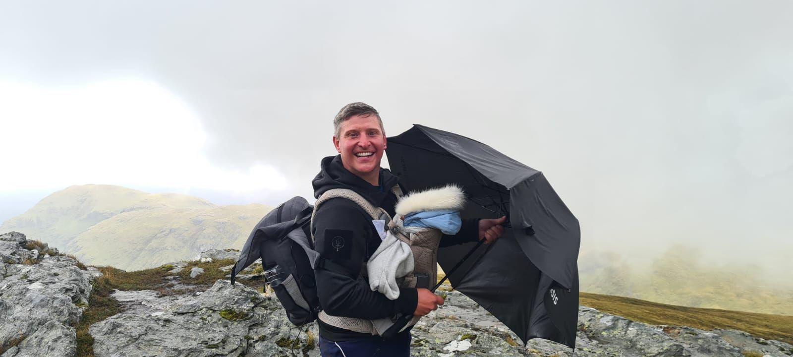 Man smiling, carrying a baby in a chest worn carrier, holding an umbrella over him, standing on top of a munro