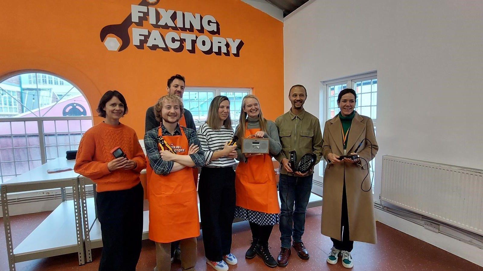 A group of people, some wearing orange aprons and holding different electronics, stand in front of an orange sign which reads: "Fixing Factory"