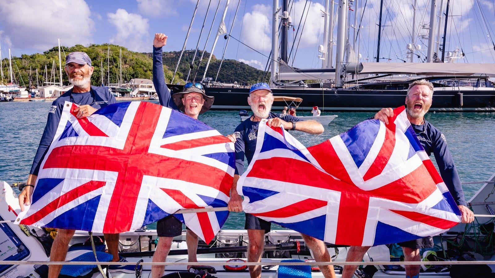 The same four men as in the first image, in similar scenery. They appear nearer to land, or may be docked. Instead of flares, they hold two Union Flags between the four of them. They are cheering and smiling wide. One man holds his arm up in triumph.