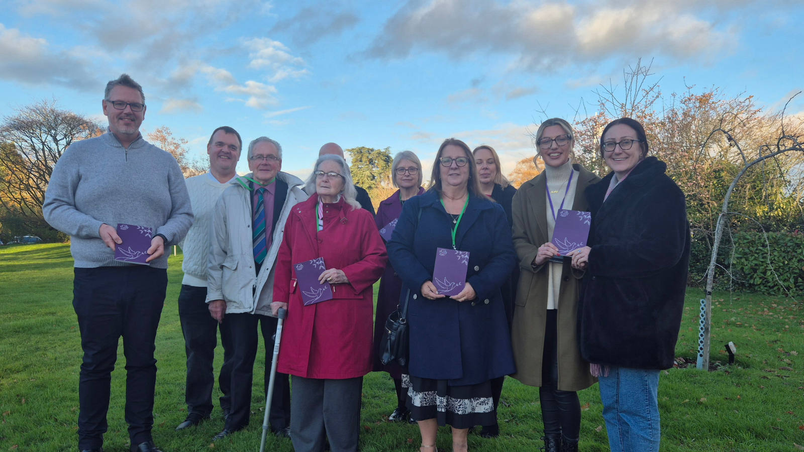 Four men standing with six women on a green grass field. They are all holding purple notebooks with white patterns on them.