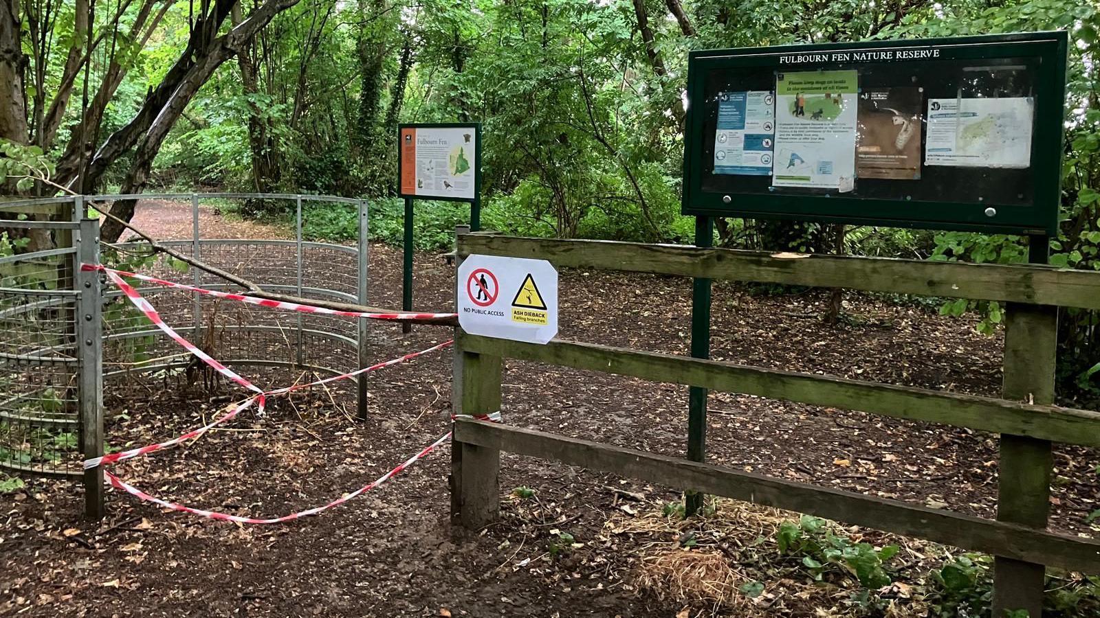 A wood, with an entrance tapped off with red tape, showing a sign for no access and a Wildlife Trust sign in the distance and another large black sign to the right. There are brown leaves and foliage on the ground and trees all around the area. 