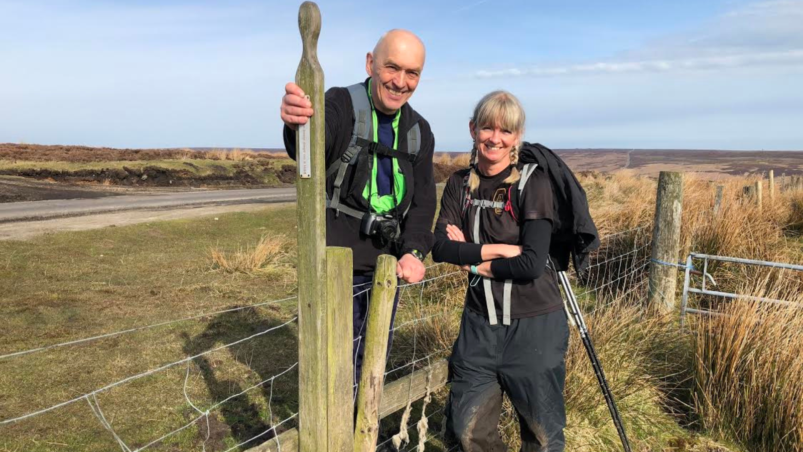 Two walkers, wearing walking gear stand by a wooden stile in the North York Moors. In the background, hills covered in heather can be seen.