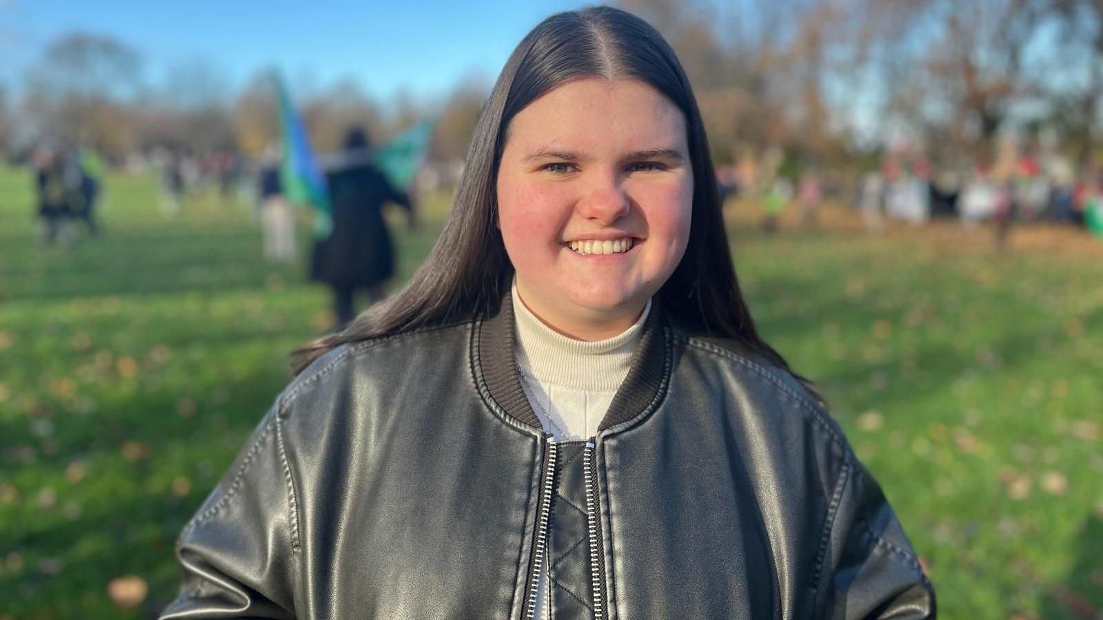 A young woman with long dark hair smiles at the camera while standing in a sunlit park. She is wearing a grey leather jacket over a light turtleneck. People and trees are blurred in the background.