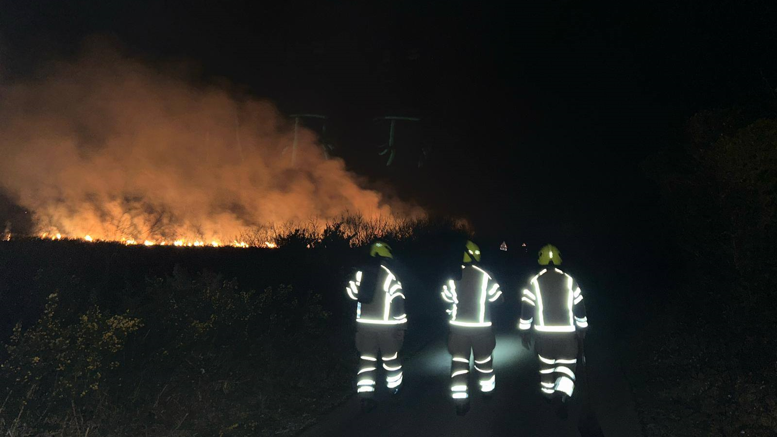 Three firefighters walking towards a blaze in the dark. Their uniforms are being lit by the flash of a camera. 