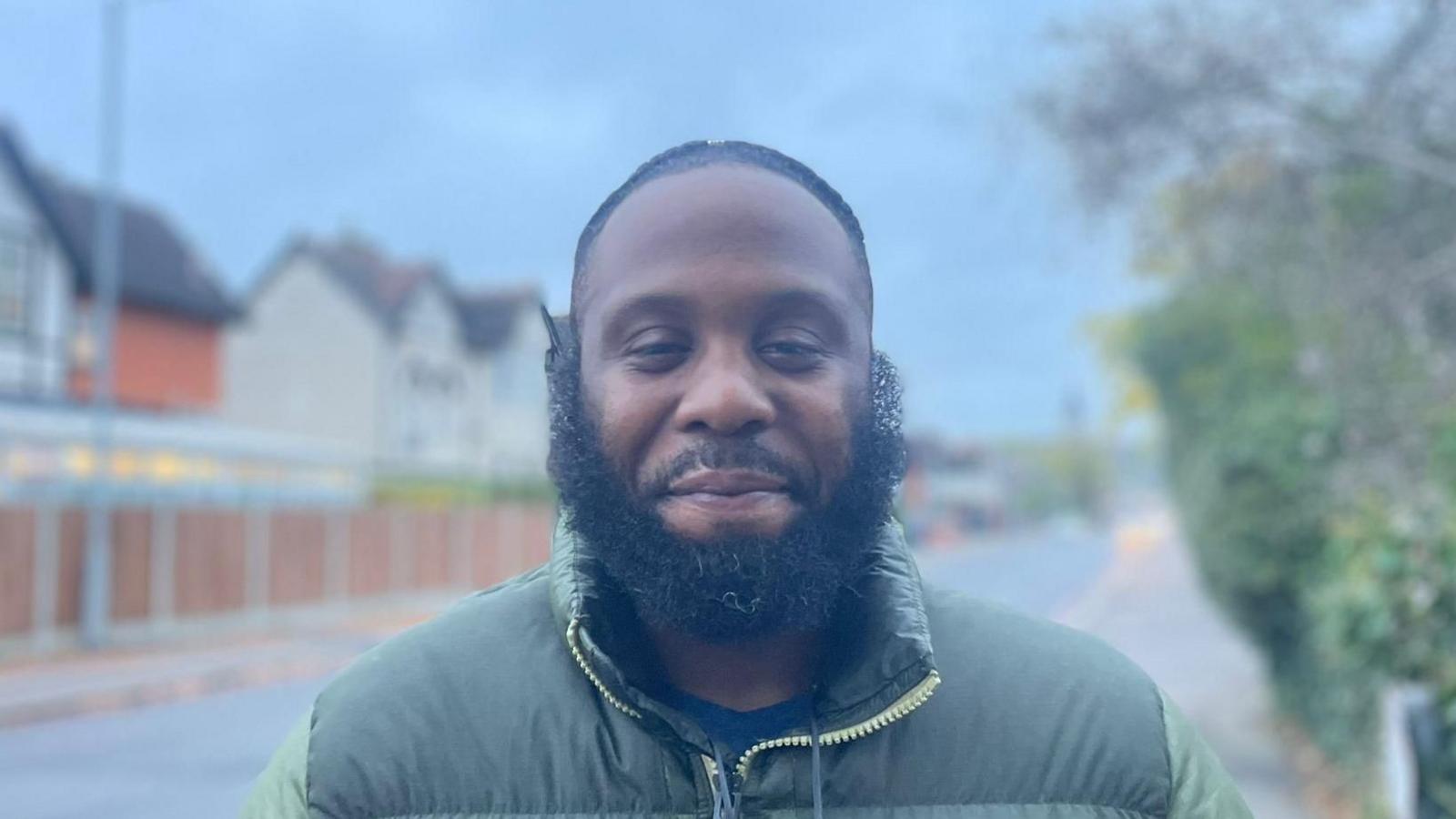 A man is smiling at the camera. He has short dark hair and a full dark beard. He is wearing a green jacket and is standing on a road with houses, a fence and greenery behind him.