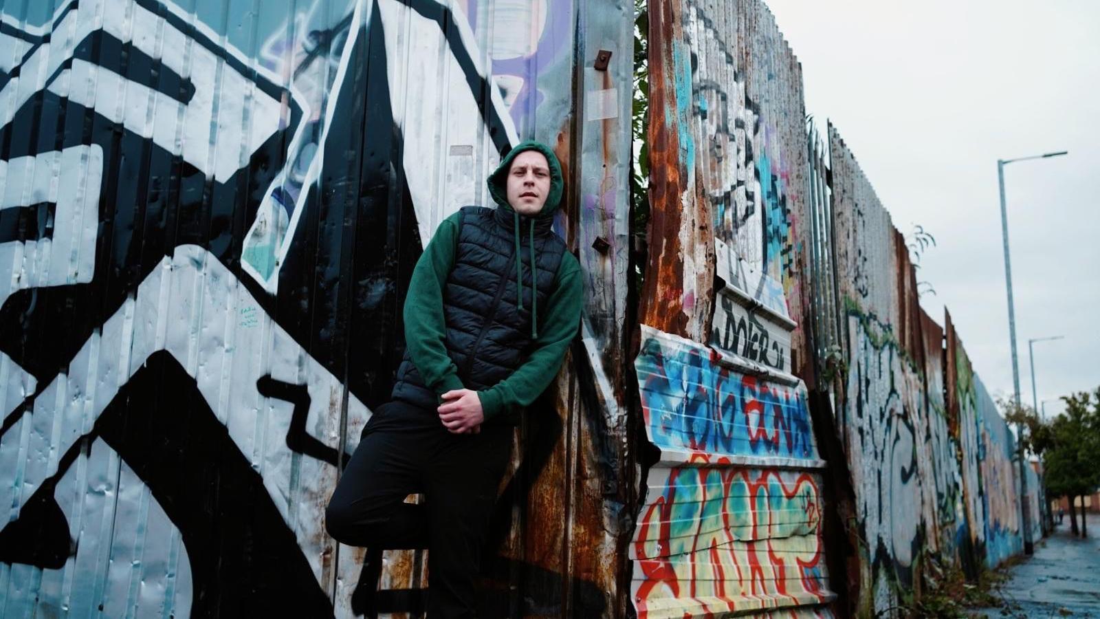 Young Spencer, wearing a green hoodie with a black, puffer gilet. He is leaning against a corrugated metal fence with graffiti on it.