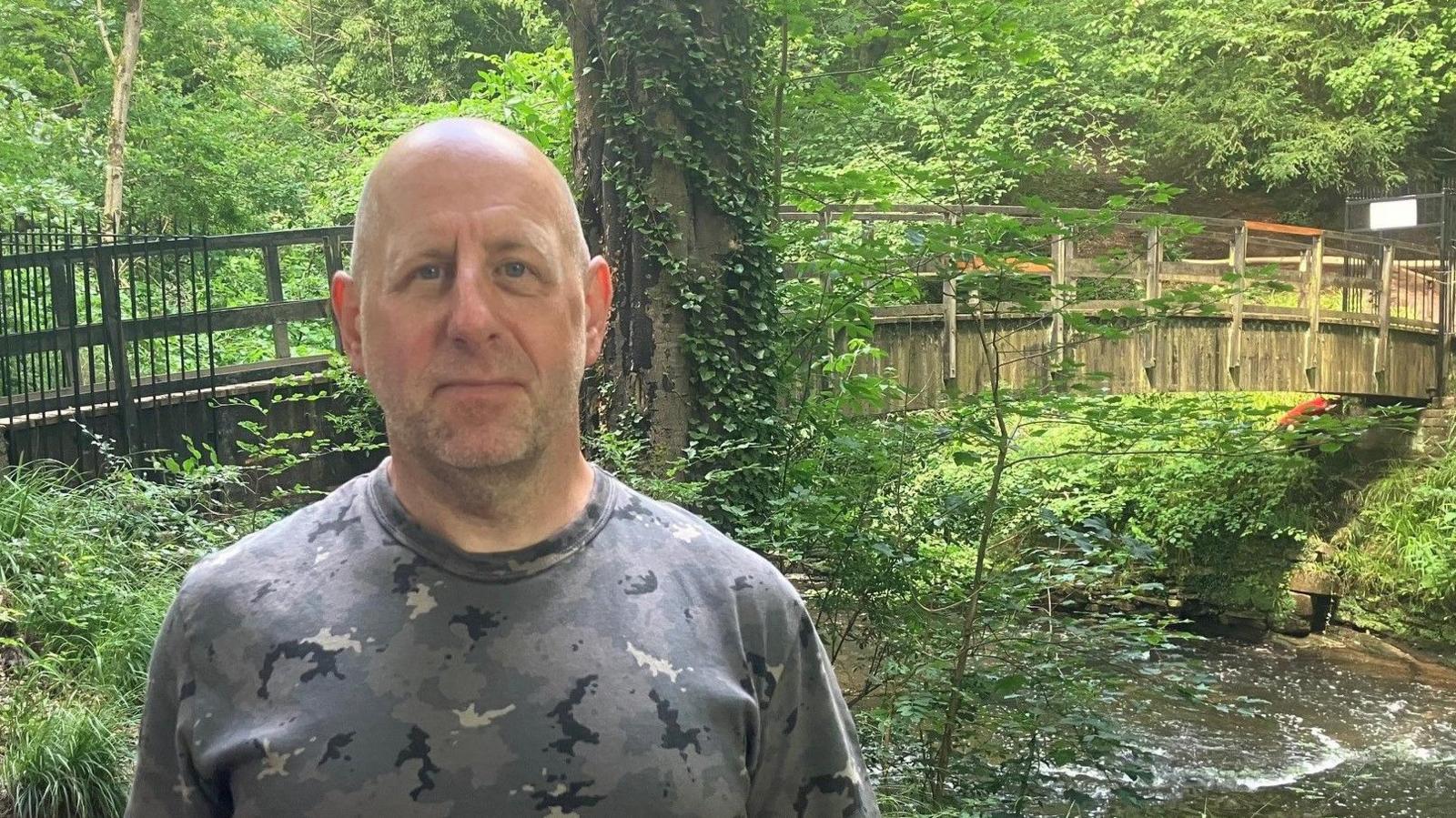 Photograph of Reece Sanford from Northern Moor in Wythenshawe in front of the Giant's Castle Bridge in Styal Woods. He is a bald man wearing a grey, black and white camouflage-style round-necked tshirt. The River Bollin flows under the wooden bridge.