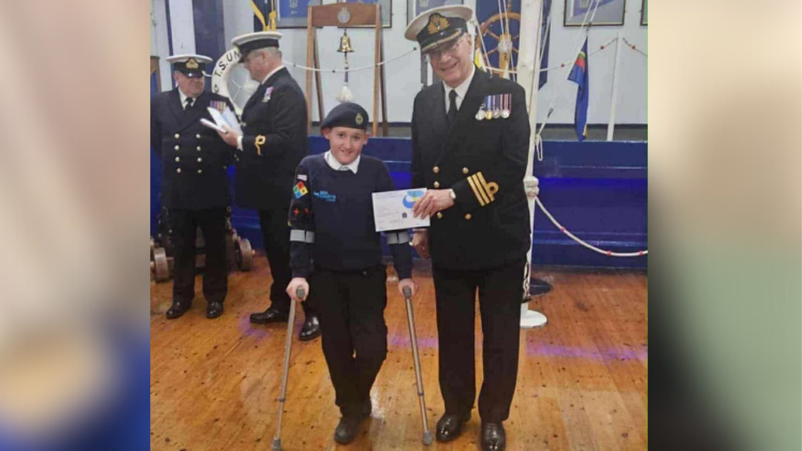 Archie is presented an award at Filey Sea Cadets by an older man in full naval uniform - white hat, blue suit with gold buttons and a gold trim. Archie wears a white shirt, navy blue jumper and trousers and a navy beret. He uses crutches.