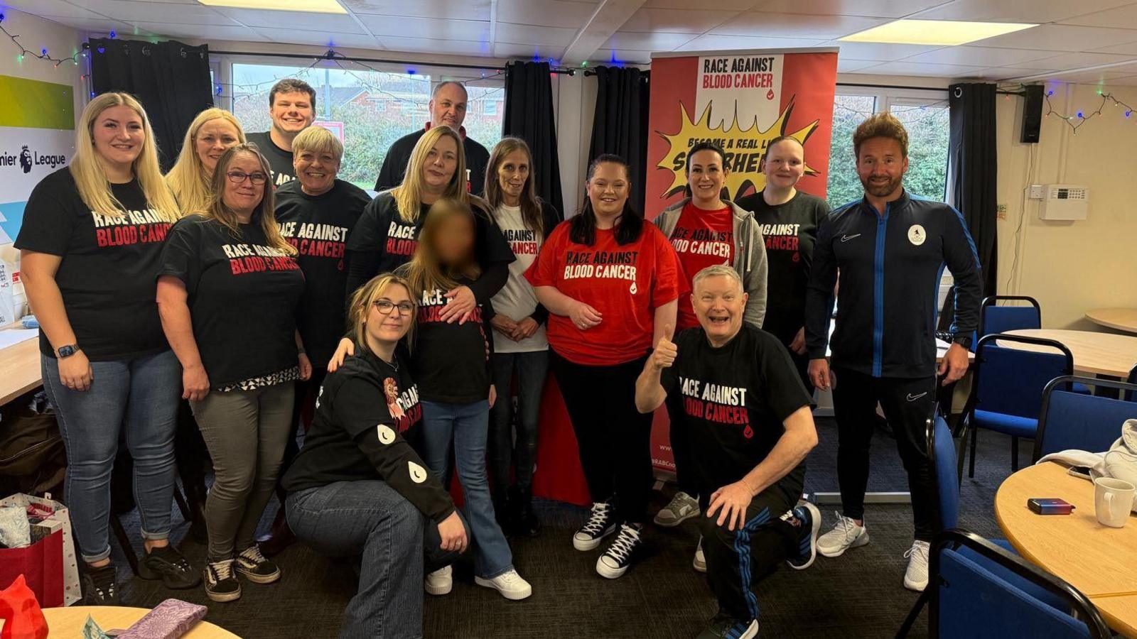 Fifteen people, men and women are standing in a room together in front of a red banner that reads "Race Against Blood Cancer". They are all wearing black or red t-shirts with the same lettering on them.