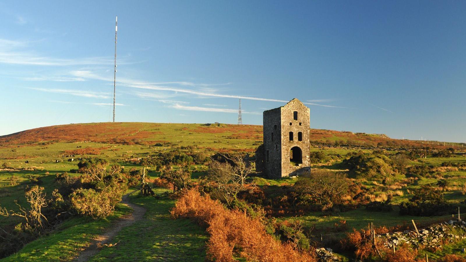 An old tin mine building in a Cornish field on a sunny day. The granite building is surrounded by shrubs and other plants. A large telecommunications pole is in the distance along with a telecommunications tower.