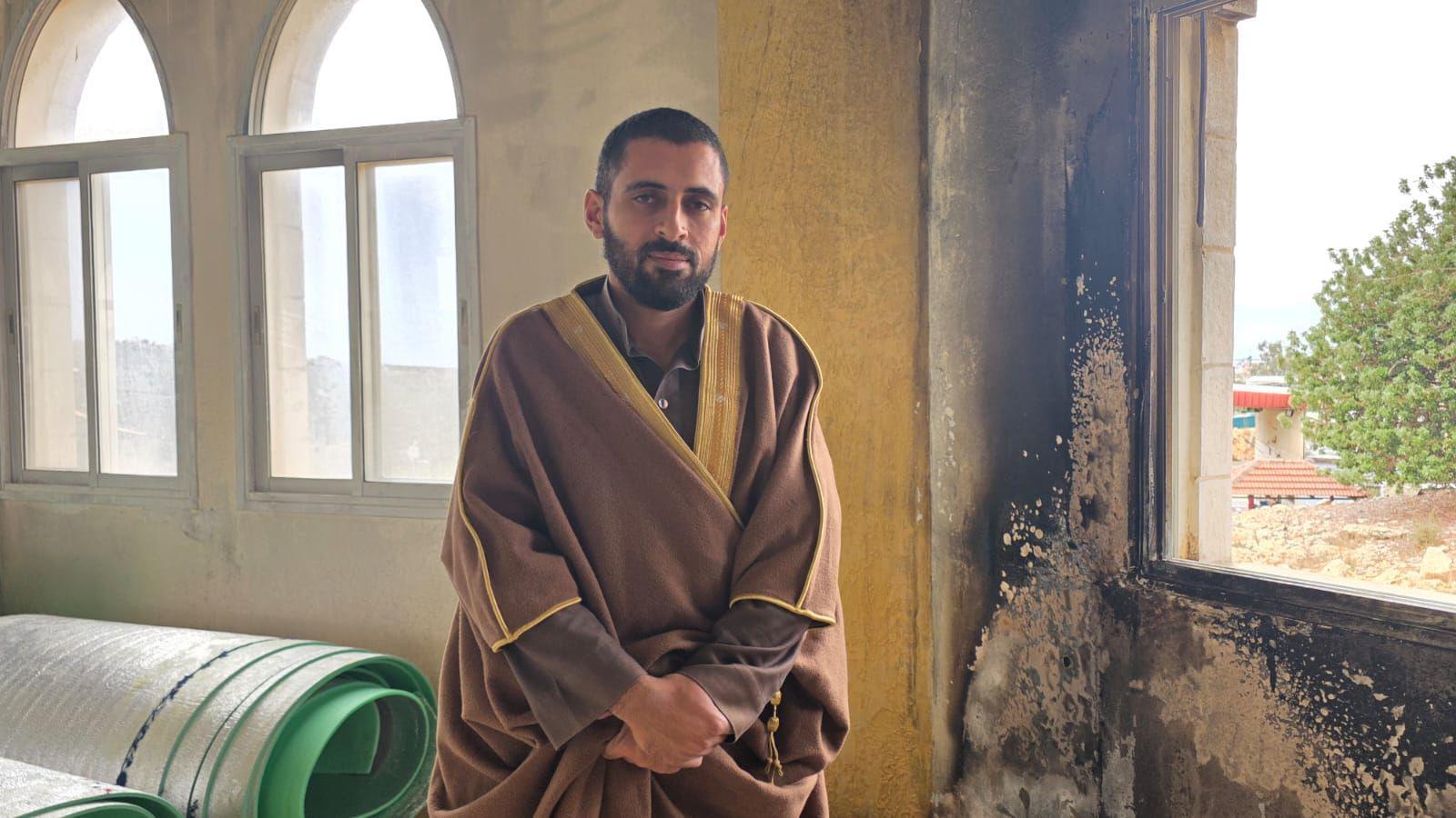 Imam Ahmad Salman. He is wearing a brown robe with yellow detail. He is standing next to a burnt wall where the paint has been blackened. The window next to it is empty of glass.