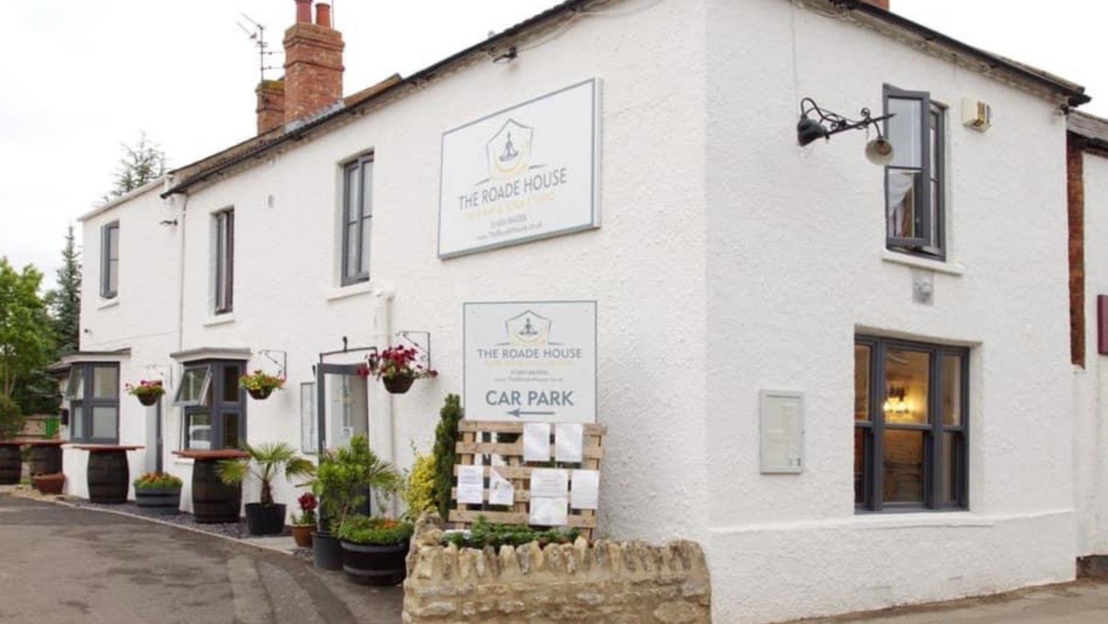 An exterior photo of a white cottage with lots of shrubs and barrels outside. There is a sign for the car park on the edge of the building. 