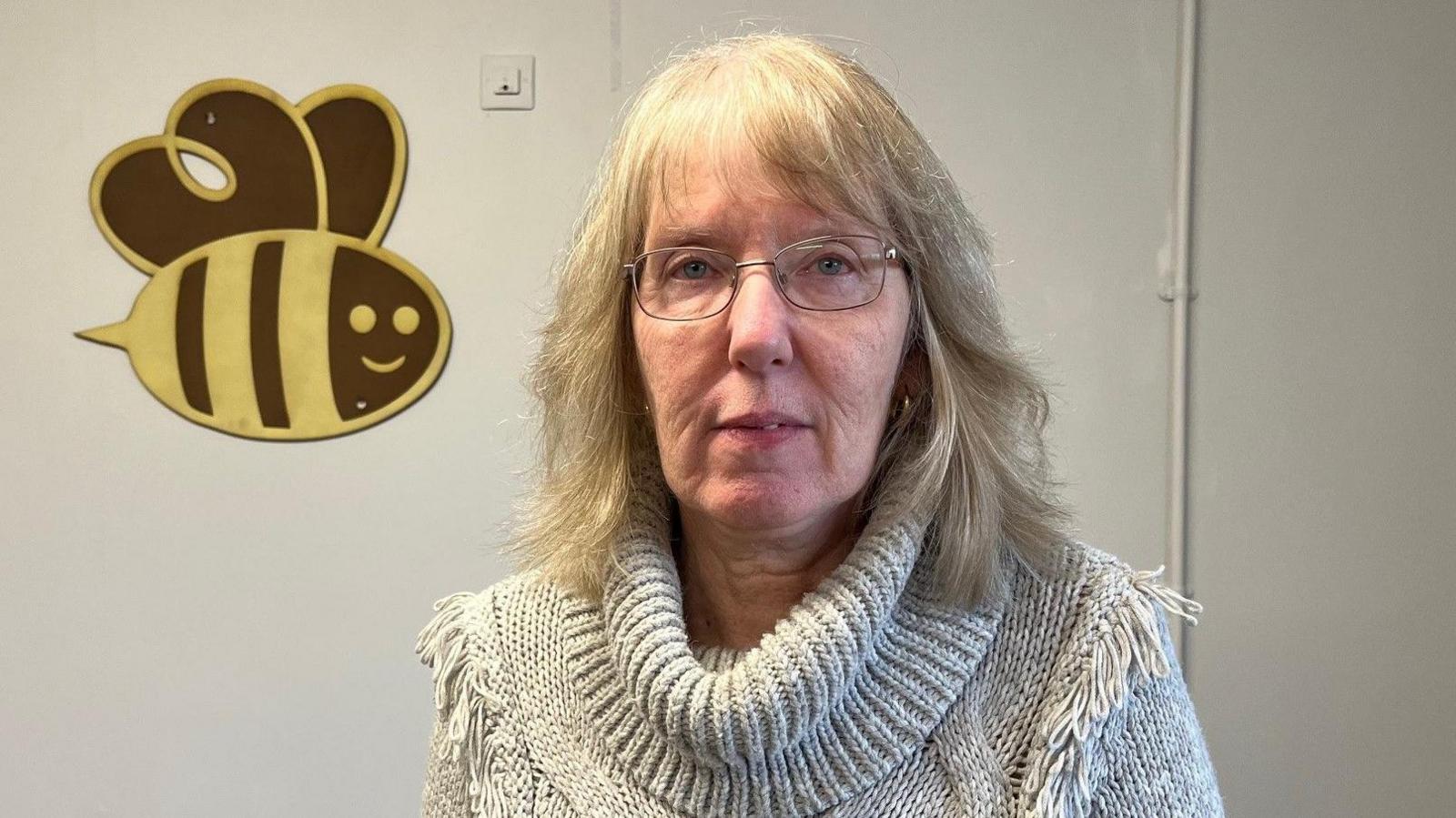 Pamela Hodge, who has blonde hair and is wearing a cream roll-neck jumper, stands in front of a white wall decorated with a picture of a bumble bee