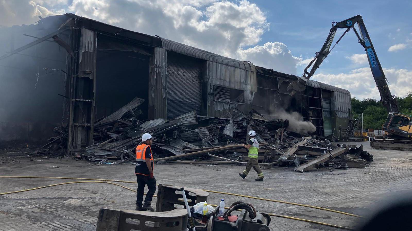 Burn out remains of a waste station, showing the building smoke damaged, parts of it torn down, two workers on the site and a crane to the right.
