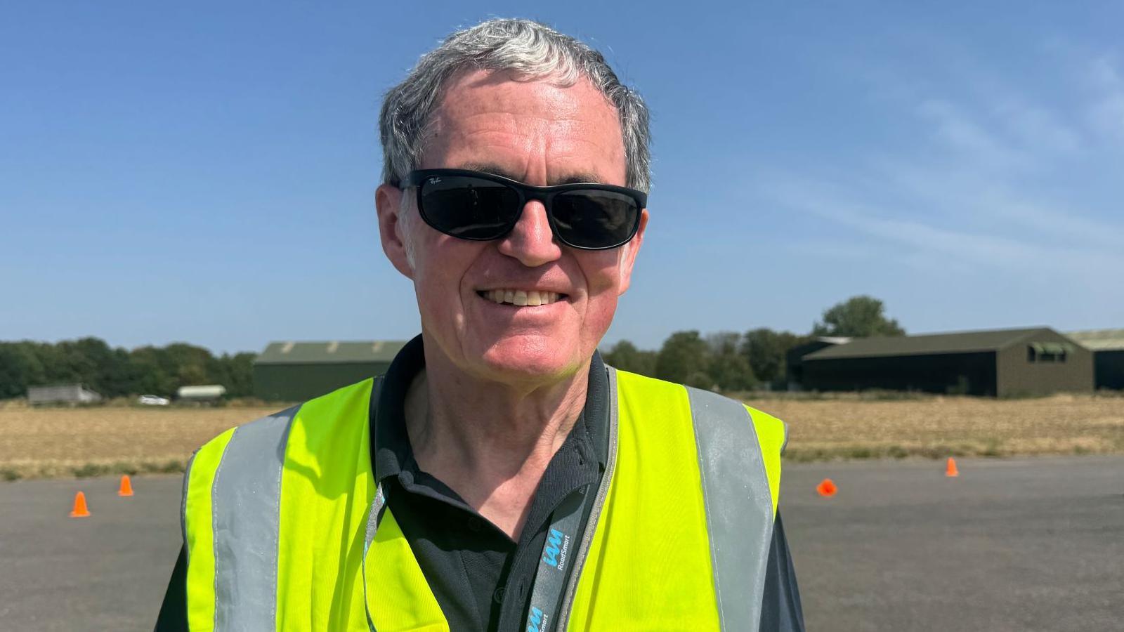 A man with short grey hair, who wears sunglasses and a hi-vis vest, stands on an airfield. The tarmac is lined with orange cones.
