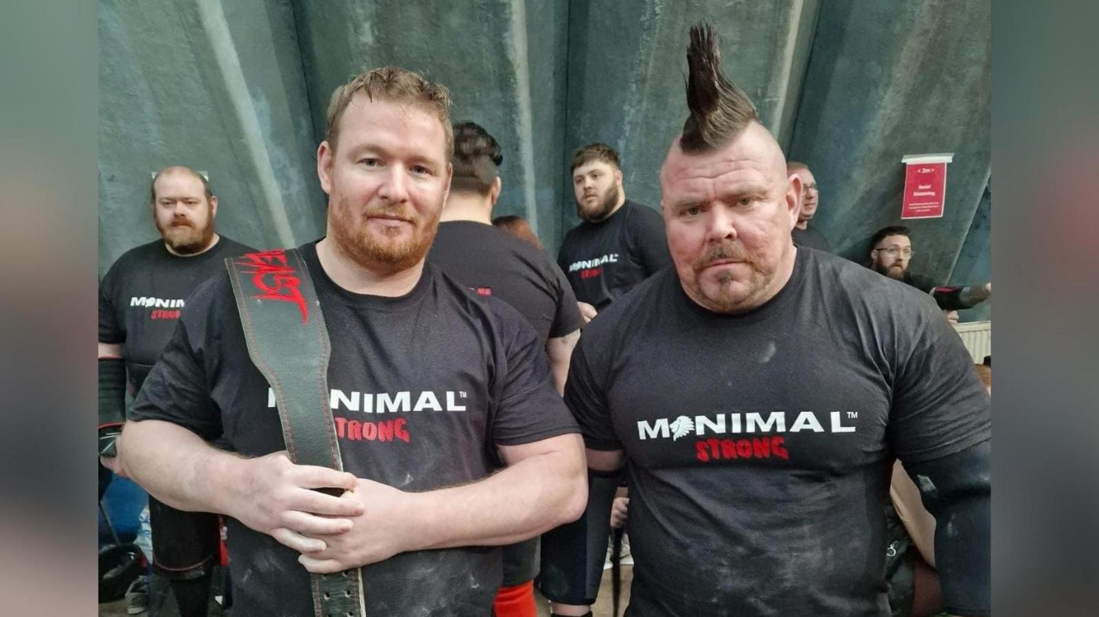 Two men in matching exercise clothing pose with a weight belt. 