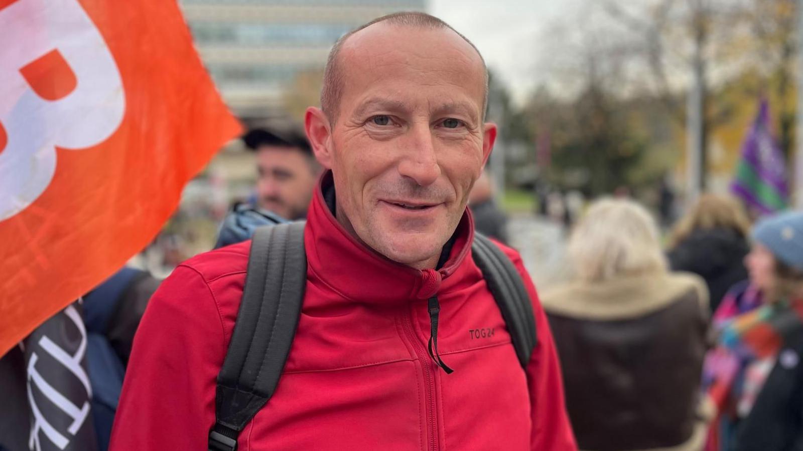 A man with shaved head and a red jacket stands with an orange flag