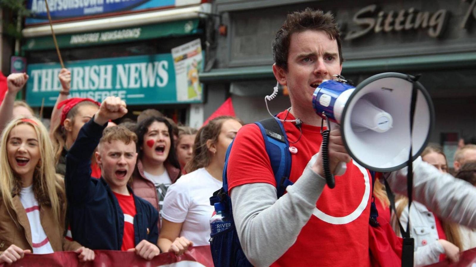 Dr Pádraig Ó Tiarnaigh in red tshirt holding a megaphone with a crowd behind him cheering at an Irish language rally.