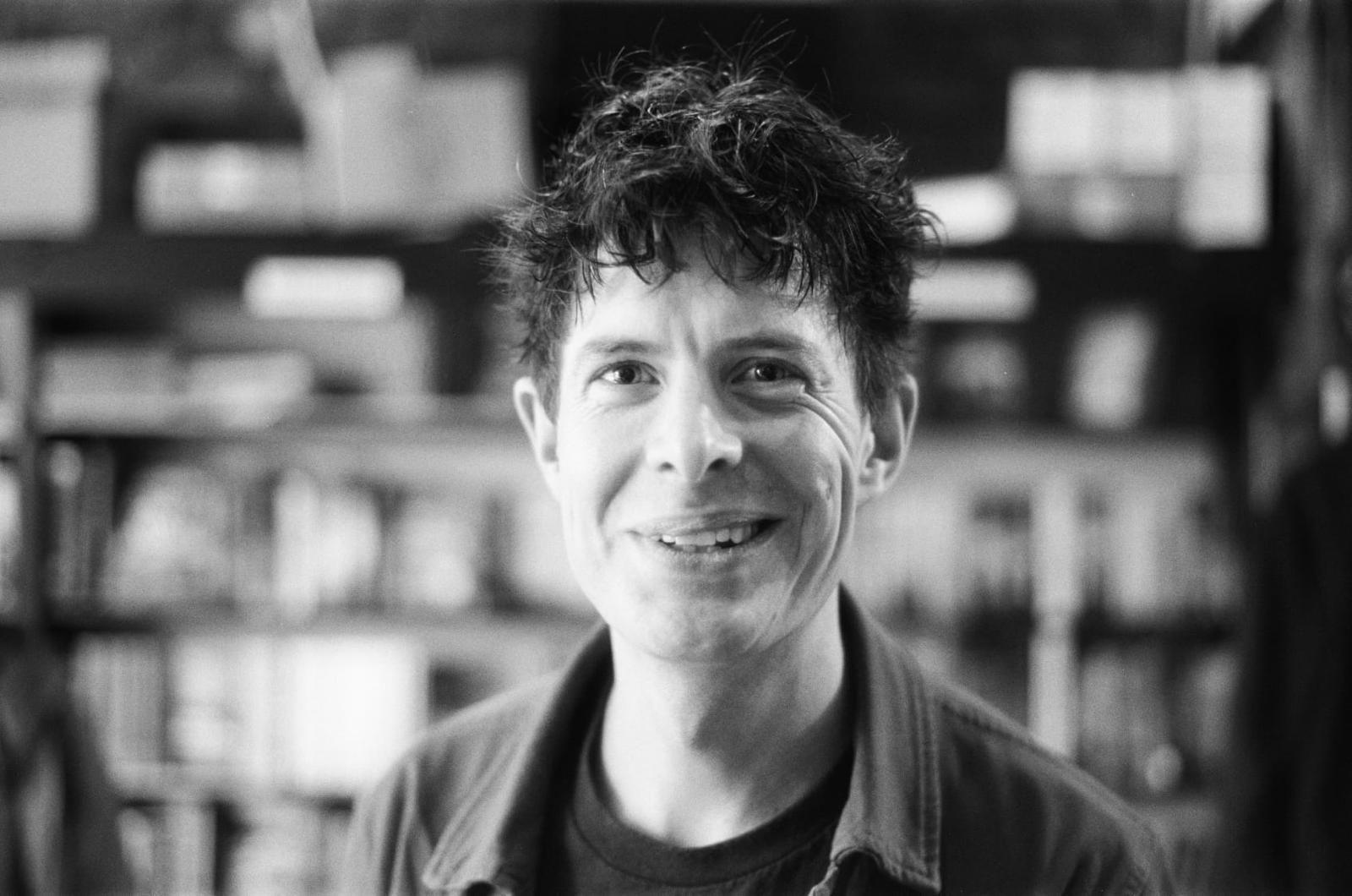A black and white image of a man smiling at the camera. He has dark hair and is wearing an open shirt.