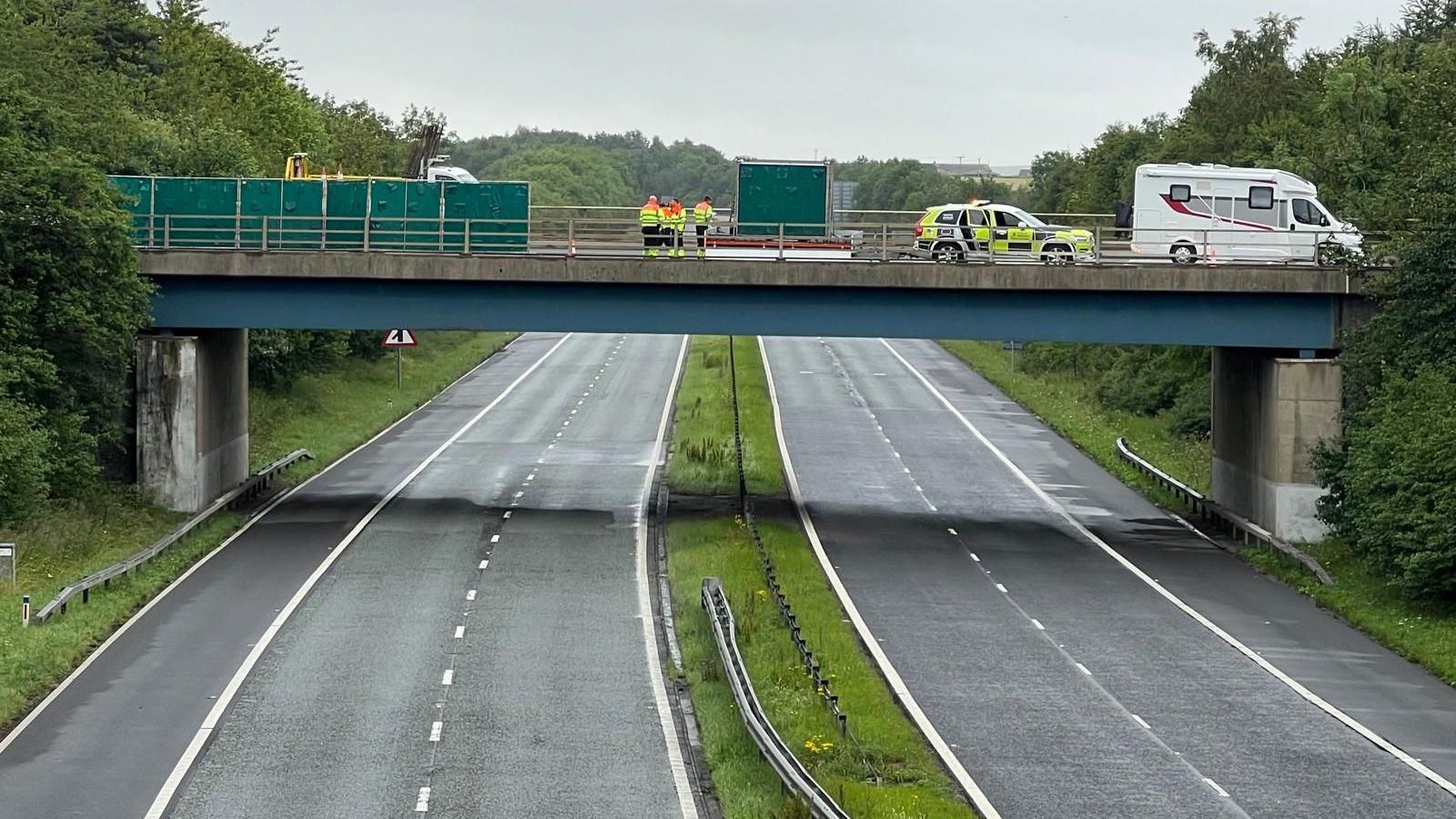 The empty A1 which is a dual carriageway with a central reservation. There is a bridge above the road with emergency service workers in hi-vis standing on it looking down. There is also a police car and caravan parked.