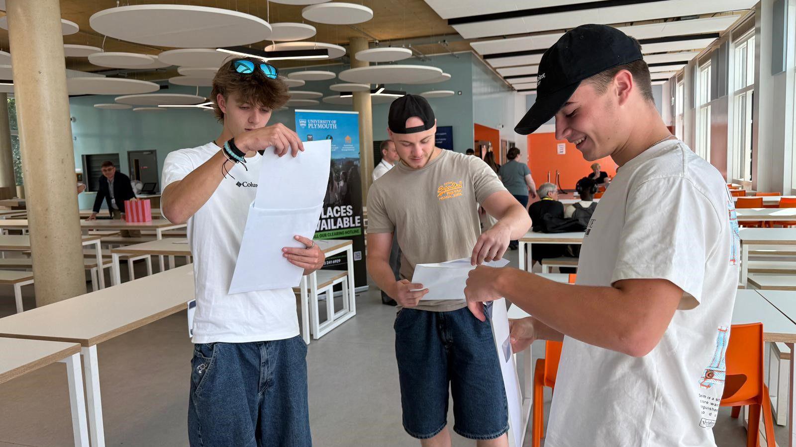 Three young men are standing in their college canteen, holding their results papers and looking down at them. From left to right, the first man has mid length dark blonde hair, wears a white short sleeved t-shirt with baggy indigo denim trousers. He has blue reflective sunglasses on his head. In the middle, the young man is bald and wears a black cap backwards with a beige short sleeved t shirt and baggy dark denim shorts. On the right, a young man with short hair wears a black cap and short sleeved white t shirt, he is smiling as he goes to open an envelope. In the background of the canteen there are a number of tables in rows and orange chair, with some people gathered in the distance.
