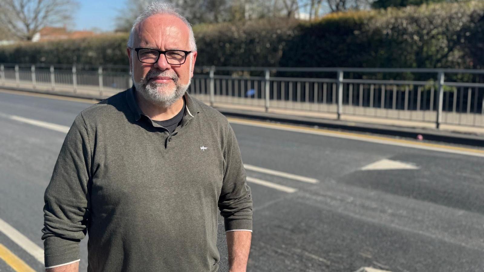 A man wearing a khaki jumper over a black T-shirt looking straight at the camera with a neutral expression. He is standing on a pavement in front of a road with newly installed speed bumps.