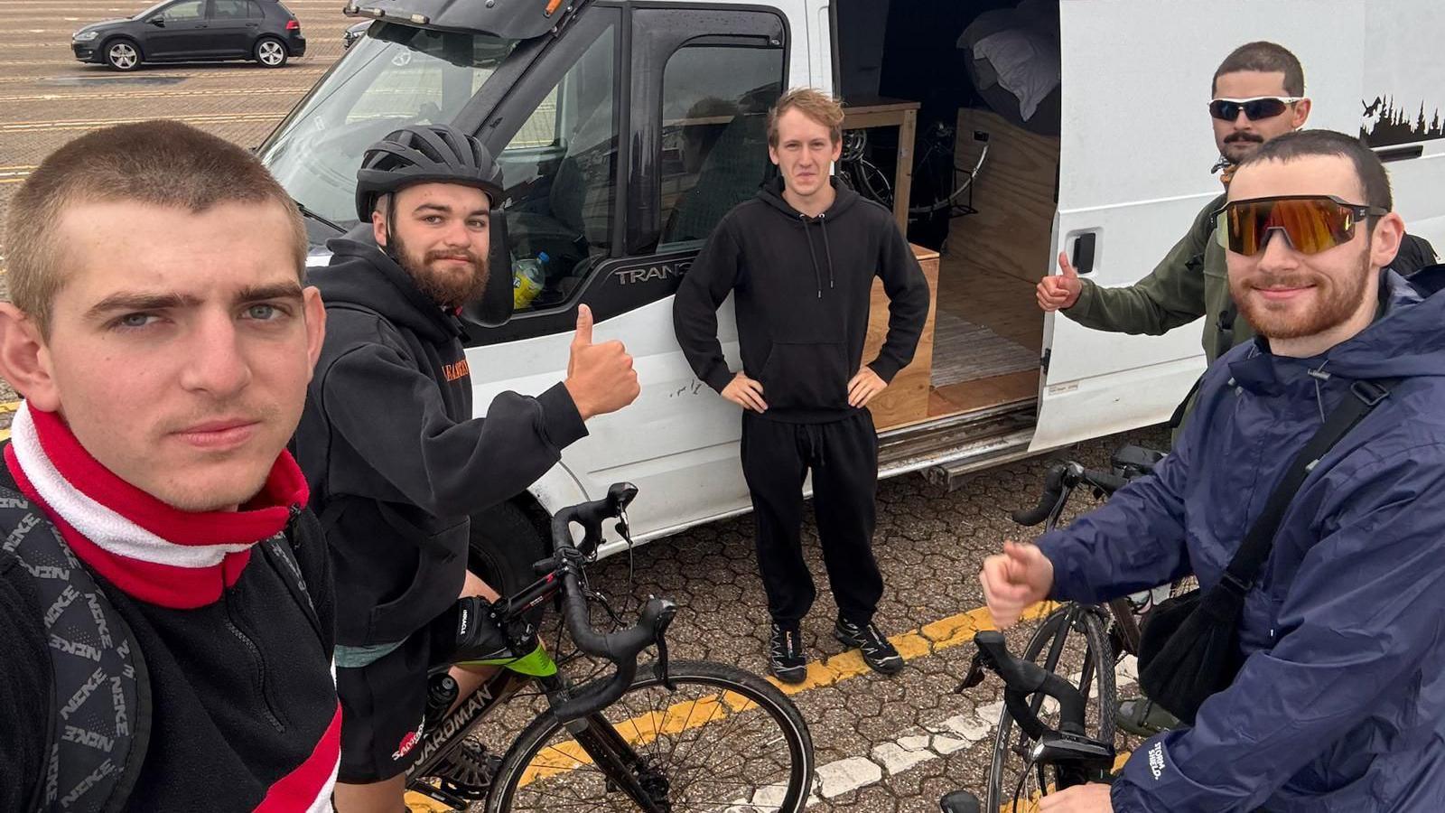 Five men, four of whom are standing with bikes. They are outdoors and are standing next to a white van.