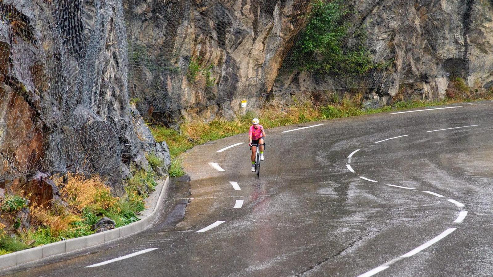 A woman cycling far away in the middle of a winding road with a rock wall and wet road