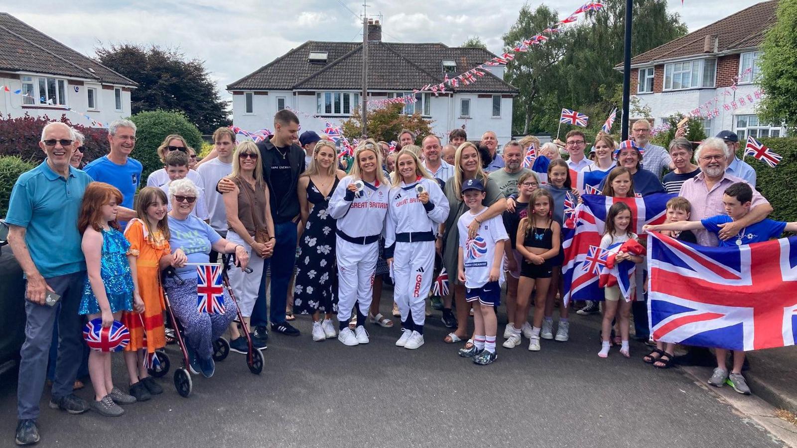 Olympic medallists Kate Shortman and Isabelle Thorpe stand in their Team GB tracksuits among a group of family and friends on their return to Bristol. People in the group are holding up Union flags and smiling at the camera, with houses in the background