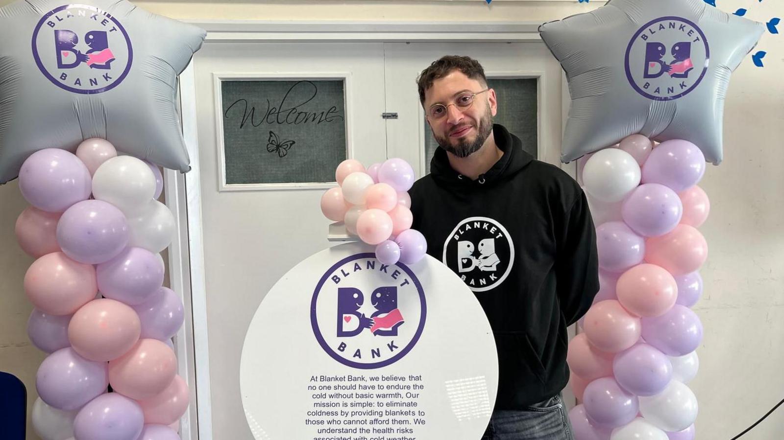 Paris Theodorou is surrounded by balloons at the launch of Blanket Bank. He has short dark hair and a dark beard and wears glasses. He has a black hoodie on with the words "Blanket Bank" in a circle on it and stands behind a circle with writing about the bank on it. Either side are swirls of lilac, pink and white balloons and he stands in front of white doors.