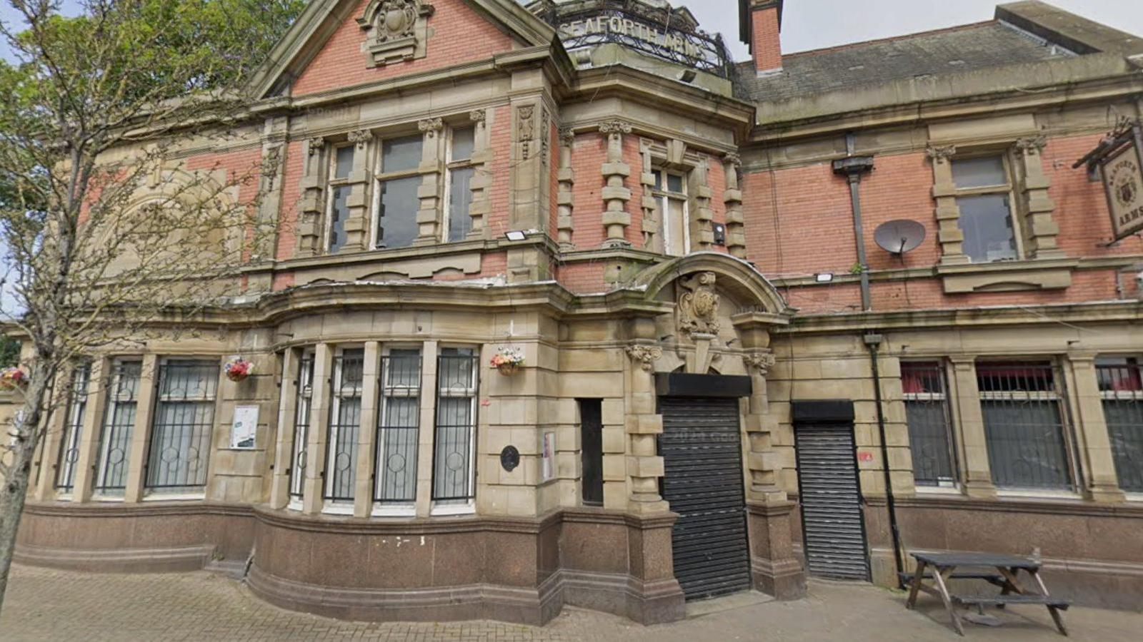 The outside of a red brick and sandstone pub building. A tree grows through a pavement in front of the two-storey building, which can be seen on an overcast today.