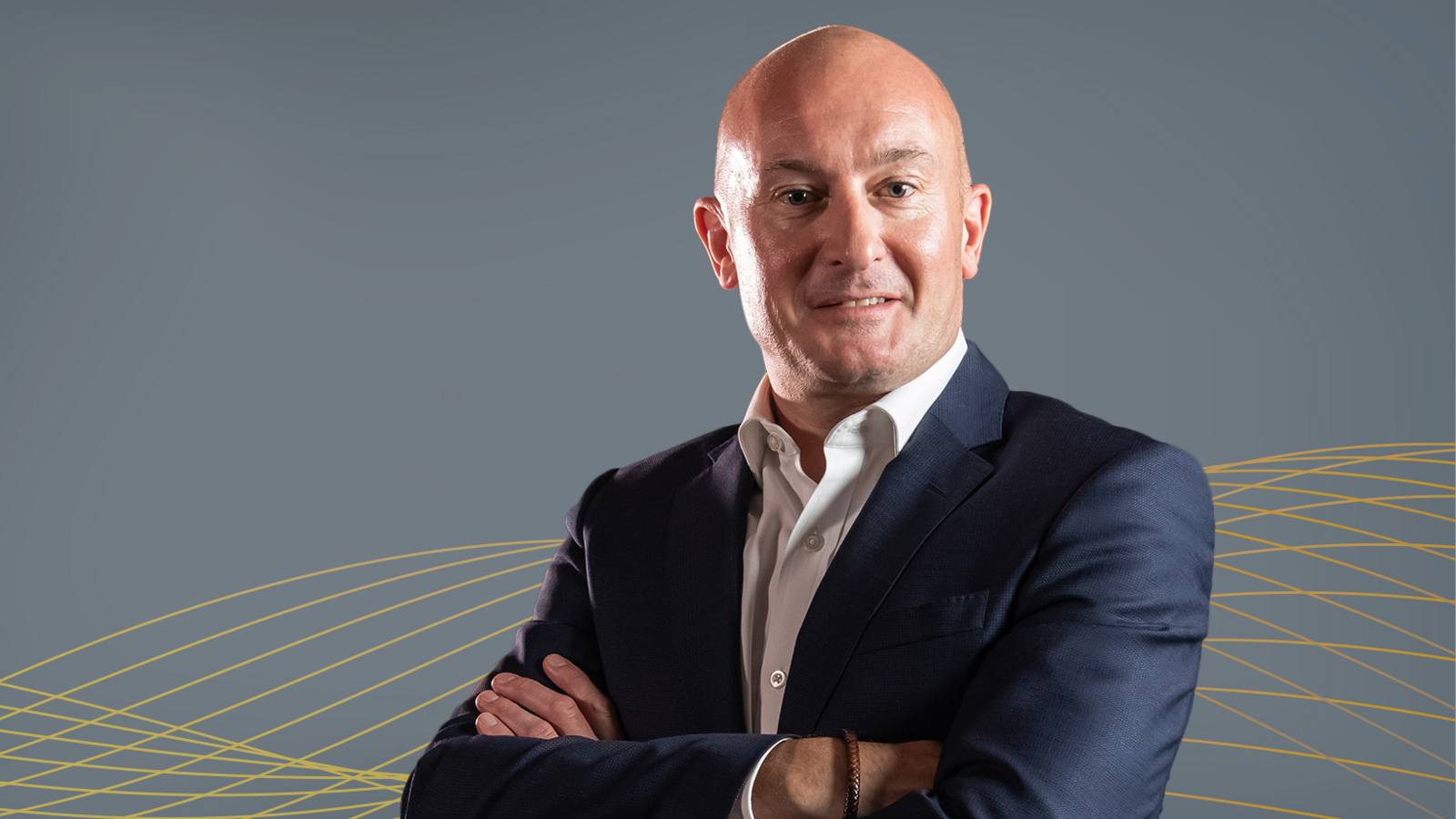 David Larssen is a bald man in a blue suit and open-necked white shirt. He is standing with his arms folded, turned to his left. The backdrop is a grey wall with yellow patterns.