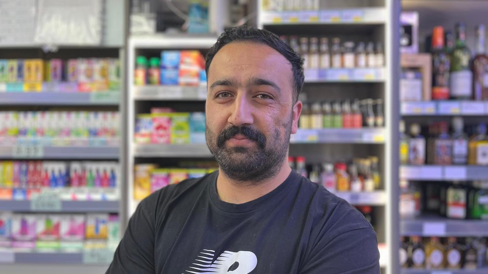 Mohammed Musazai, with black hair and a black moustache and beard, stands behind the counter of a shop with goods including spirits, vapes and medicines stocked on shelves behind him.