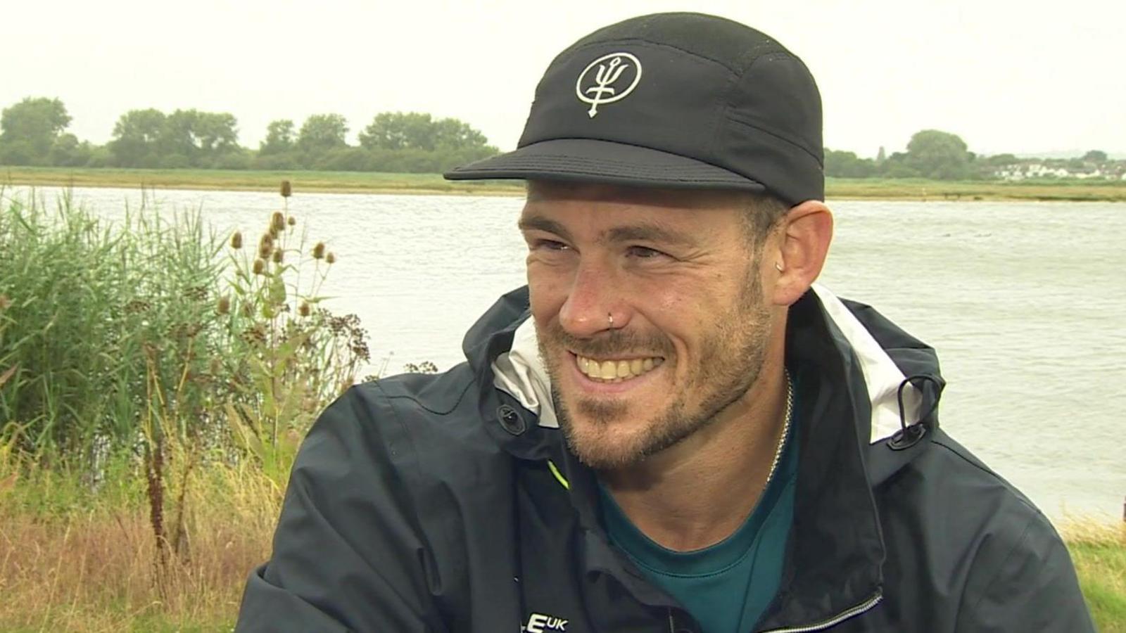 A smiling Jack Eyers with Christchurch harbour in the background. It looks like an overcast day and he is wearing a black baseball cap and a black hooded rain jacket. He has a short full beard and a nose ring.