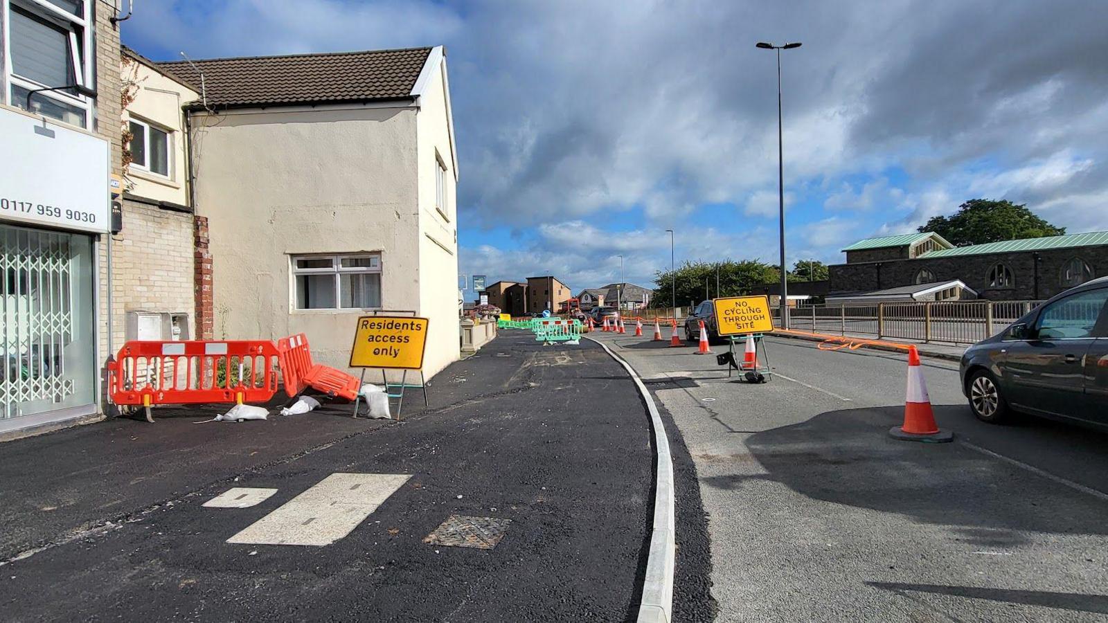 A road scene with one lane of traffic and one lane blocked off with cones and yellow signs - to the left, a newly-laid pedestrian route with orange plastic barriers