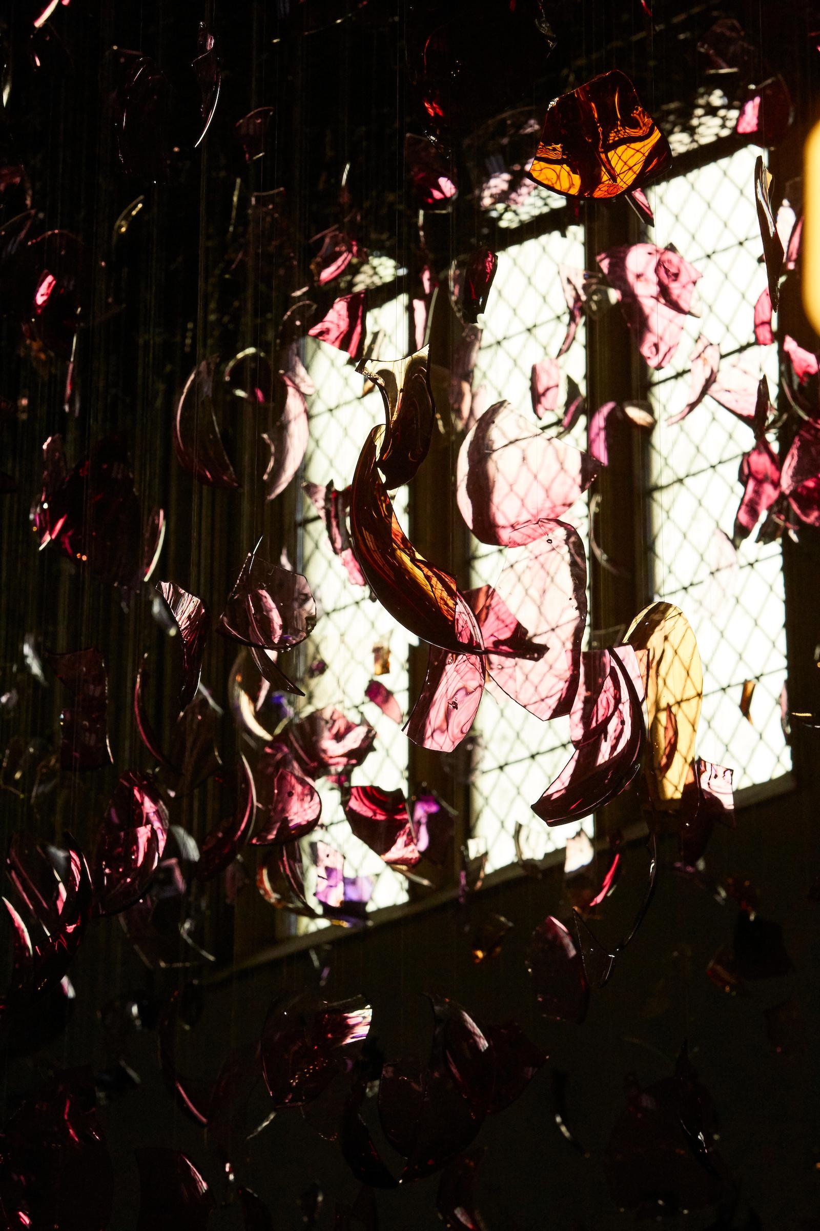 Pink, yellow, blue and purple curved glass is suspended in the air in a large church. Light shines through from a window.