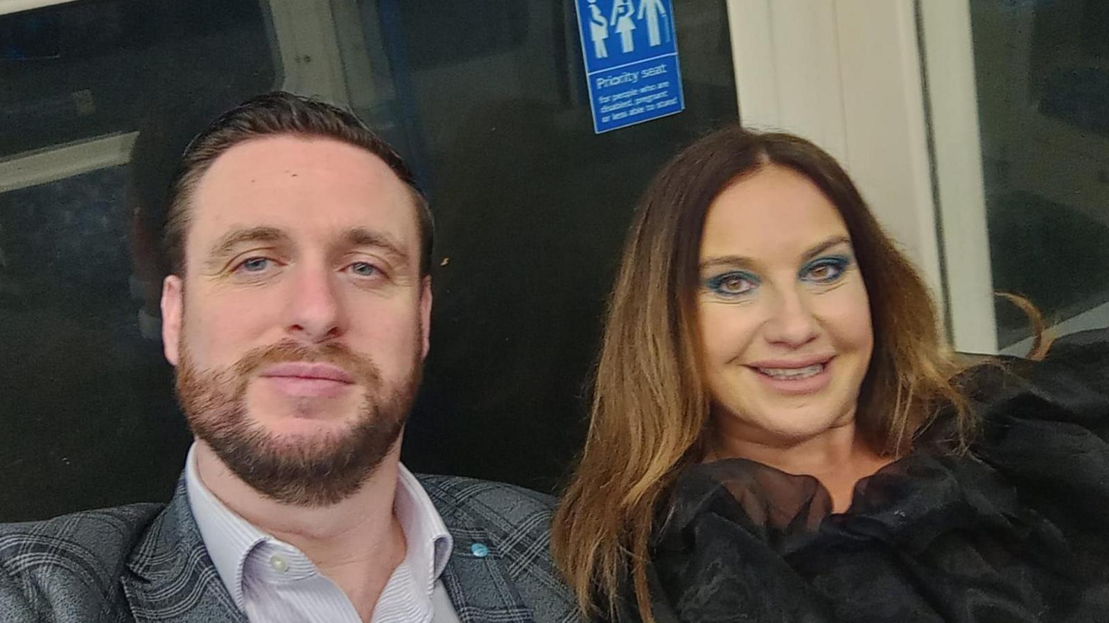 Photo of Pete and Emma Reid. Irish passengers who had planned to travel home via the storm-damaged Holyhead ferry port this Christmas. The couple are sat on the tube. Pete is wearing a checked suit jacket and stripey shirt, he has a beard and short brown hair. Emma is wearing a black chiffon blouse and has long brown hair. 