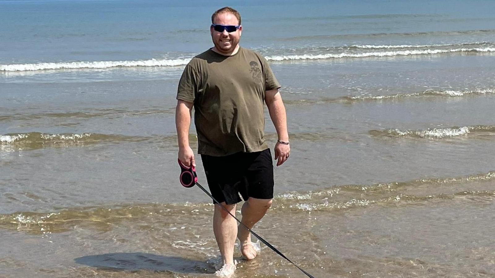 Lee Gigg wearing black shorts and a green tshirt paddling in the sea holding a dog lead