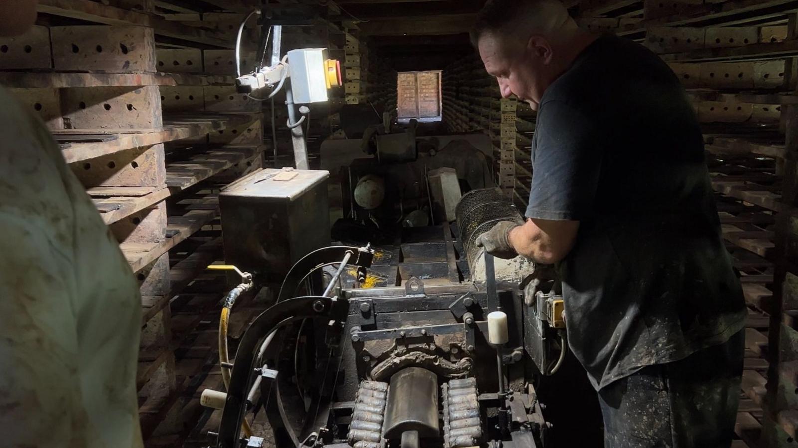 A worker is wearing a dark T-shirt and gloves operating a large piece of machinery inside a dimly lit industrial workshop. Wooden racks stacked with rectangular tiles line both sides of the narrow room, creating a corridor-like space. A single overhead work light illuminates the machine and the worker’s hands, while additional equipment is visible in the background. The scene highlights a traditional manufacturing process in a busy, enclosed workspace.