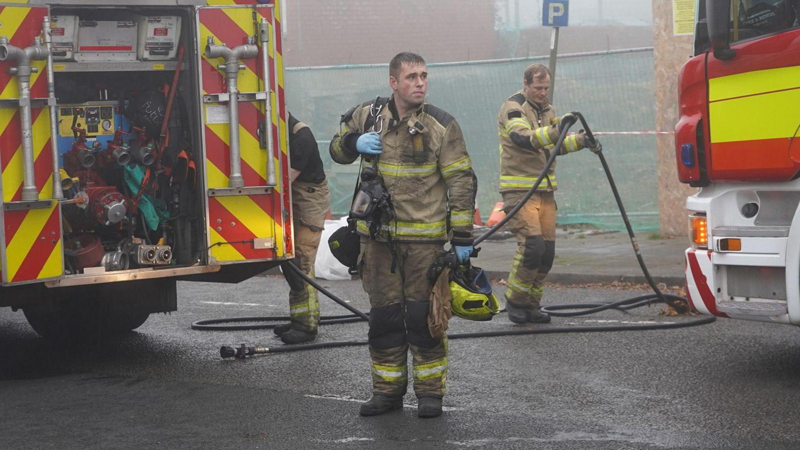 A firefighter wearing scorched yellow clothing holds a yellow helmet and mask. In the background, another firefighter moves a hose