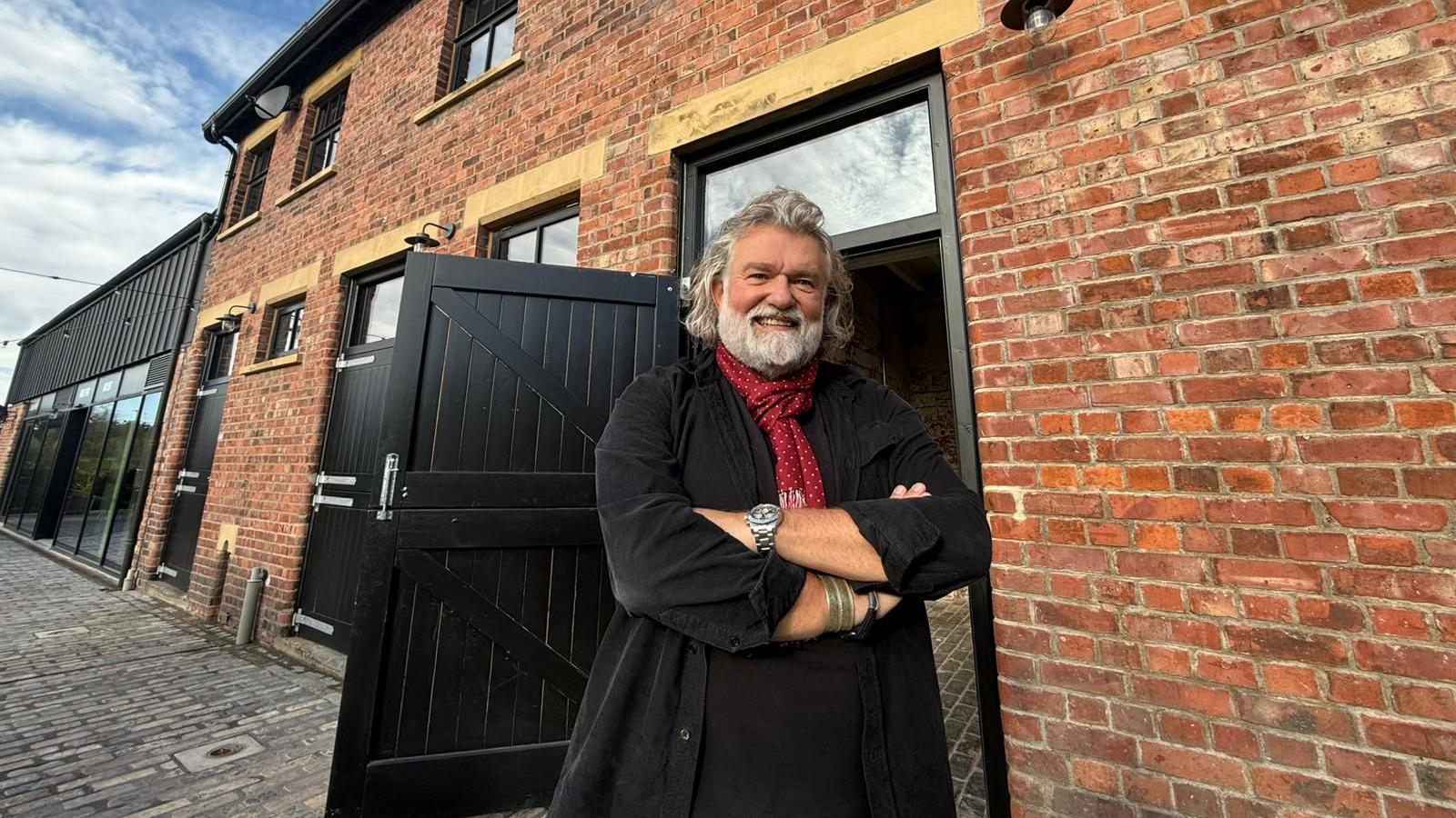 Si King standing outside the building where is new restaurant will be housed. He has grey hair and a beard and is standing with his arms crossed