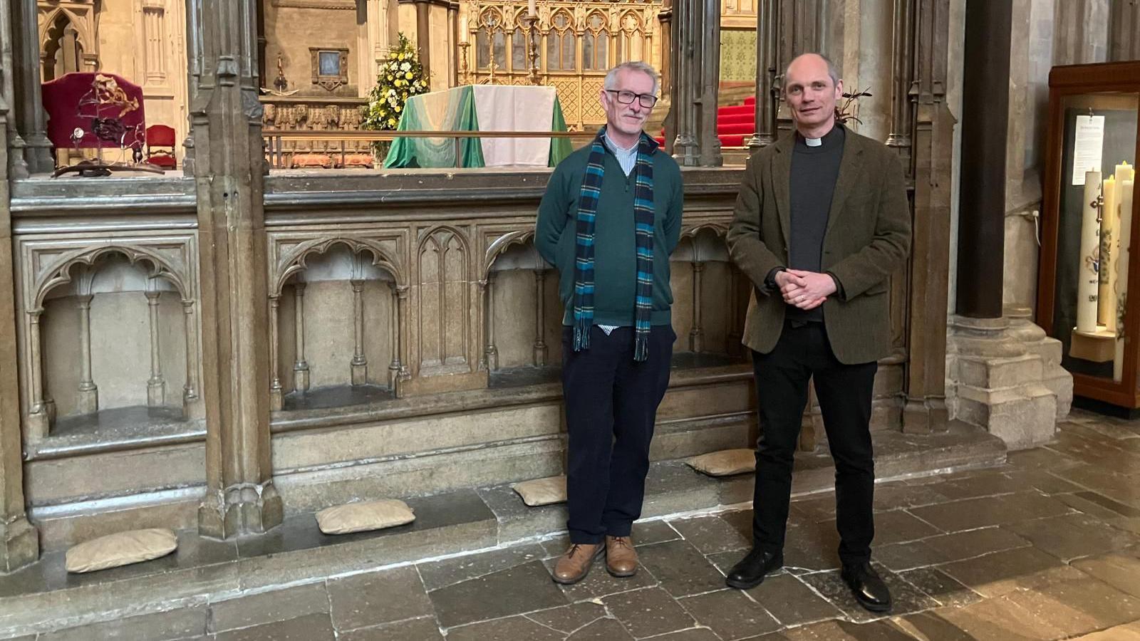 Revered Andrew Doarks and Canon Andrew Dodd standing in Canterbury Cathedral in front of Simon of Subury's tomb where his body is buried. The men are dressed in dark trousers. Andrew Doarks is wearing a green jumper and dark blue striped scarf. Andrew Dodd is wearing a dark green jacket and clerical collar. 